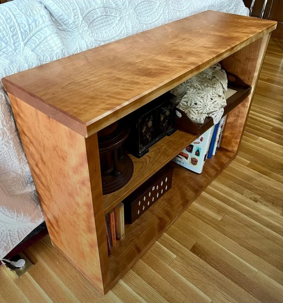 Wooden end-table with an open shelf storing books, a lace doily, and small decor items beside a couch/bed.