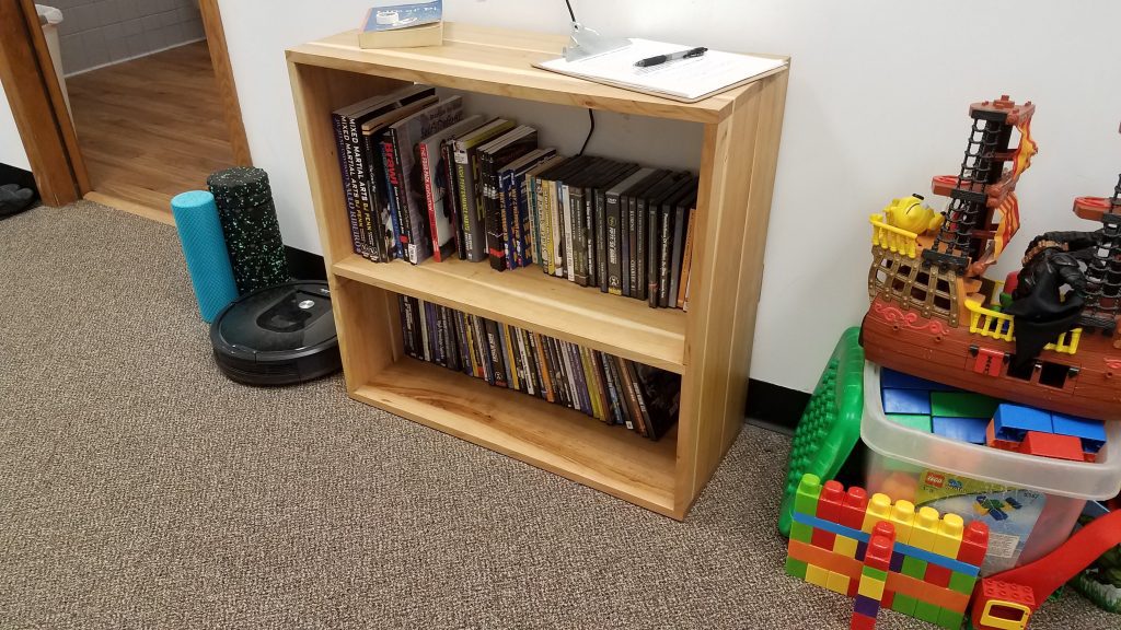 Wooden bookcase filled with books and DVDs on a carpeted floor, with yoga mats and a robotic vacuum nearby and a large toy pirate ship to the right.
