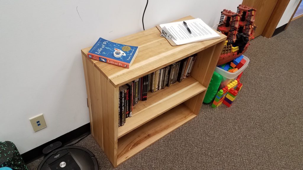 Wooden bookshelf with a blue Life of Pi book and a clipboard on top; a pirate ship model and colorful blocks sit to the right.