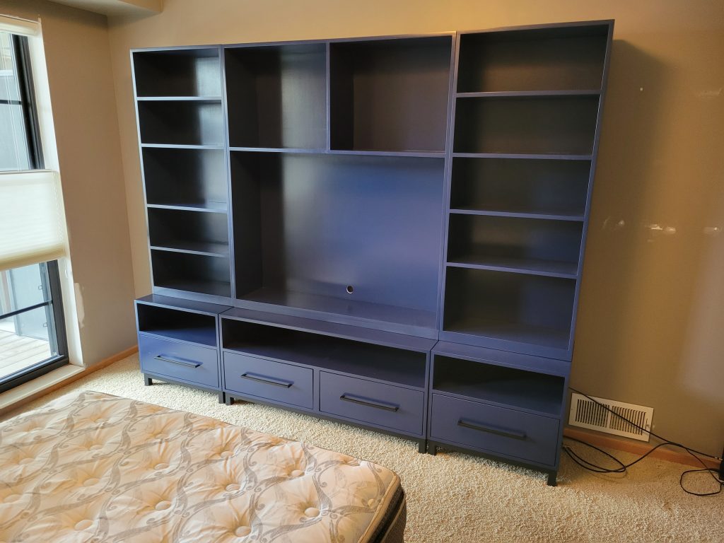 Blue modular shelving unit with multiple open cubbies and drawers, placed along a beige wall in a bedroom by a window.