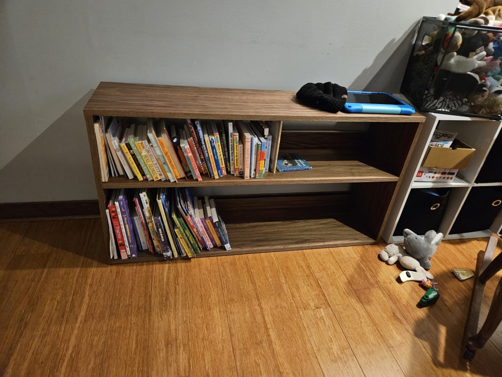 Wooden bookshelf filled with colorful children's books against a gray wall; a blue tablet with a black knit hat rests on top of the shelf.