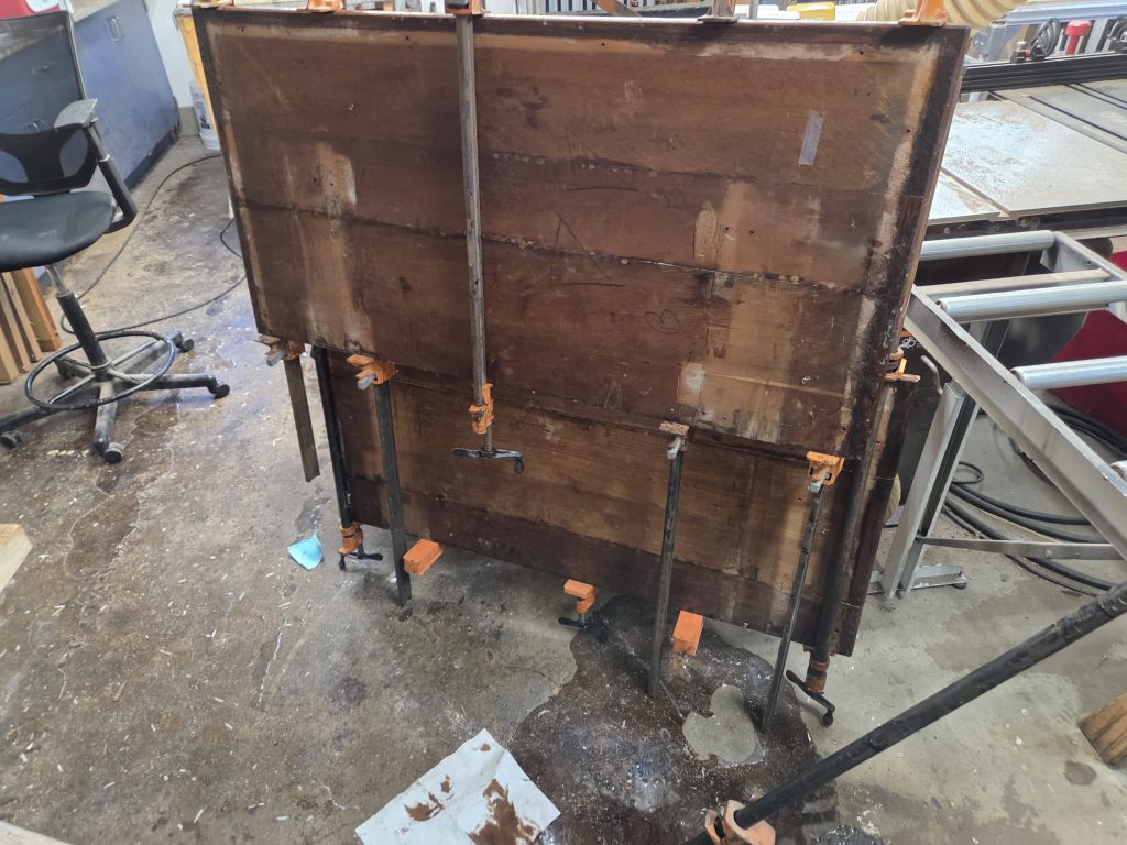 A large, weathered wooden crate mounted on adjustable metal stands with clamps in a cluttered workshop. Nearby chair, cords, and tools are visible on the concrete floor.
