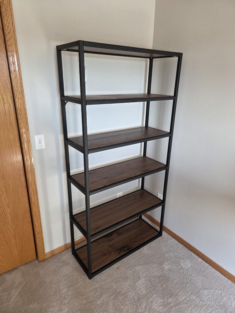 Tall black metal-framed bookshelf with five wooden shelves placed in a corner of a room next to a wooden door frame and light switch.