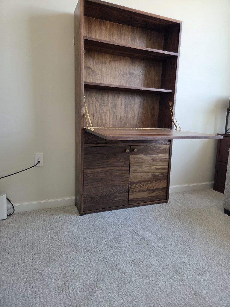 Tall dark wood secretary cabinet with open shelves and a drop-down desk surface supported by brass chains, set against a beige wall in a carpeted room.