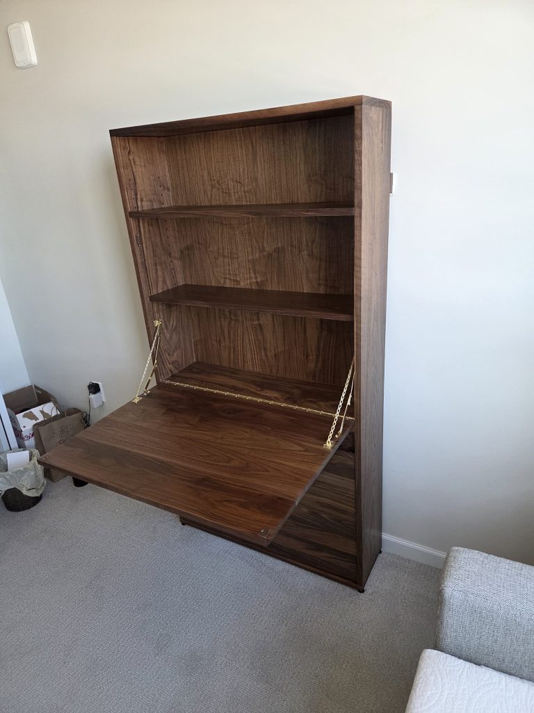 Wooden tall cabinet with a drop-down desk surface and three interior shelves, in a beige-walled room with carpet floor.