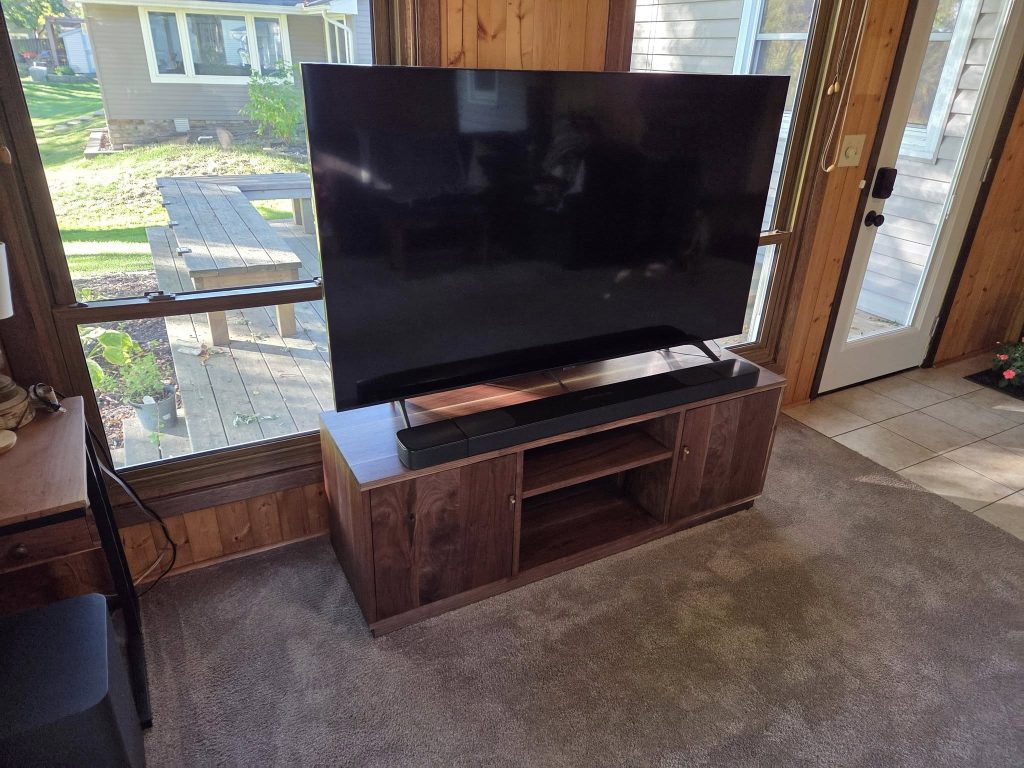 Large flat-screen TV on a wooden stand in a sunlit living room with paneled walls and a glass door outside view