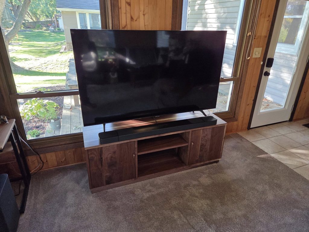 Large flat-screen TV on a wooden console in a sunlit room, with green backyard visible through the windows behind it.