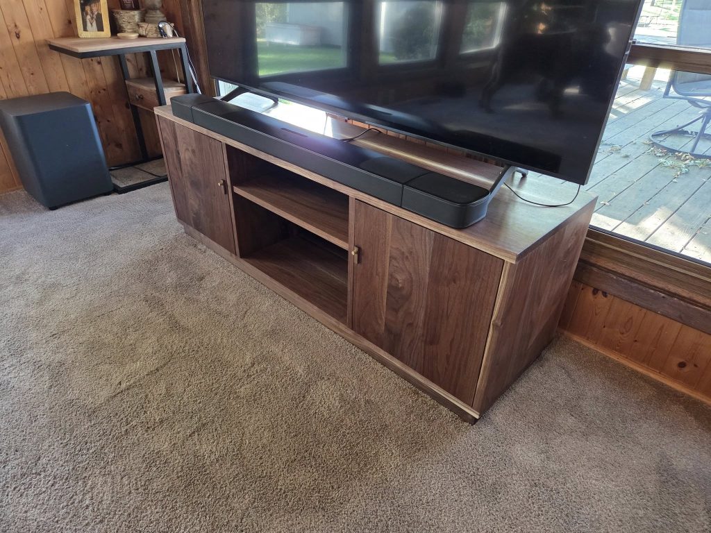 Large flat-screen TV on a wooden stand with a black soundbar and subwoofer in a wood-paneled living room.