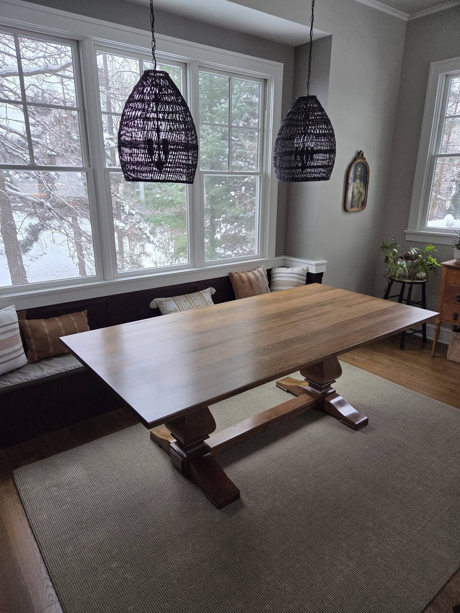 A dining area with a large wooden table, dark bench seating, and two woven pendant lights hanging from the ceiling; snowy windows outside and a small plant nook nearby.