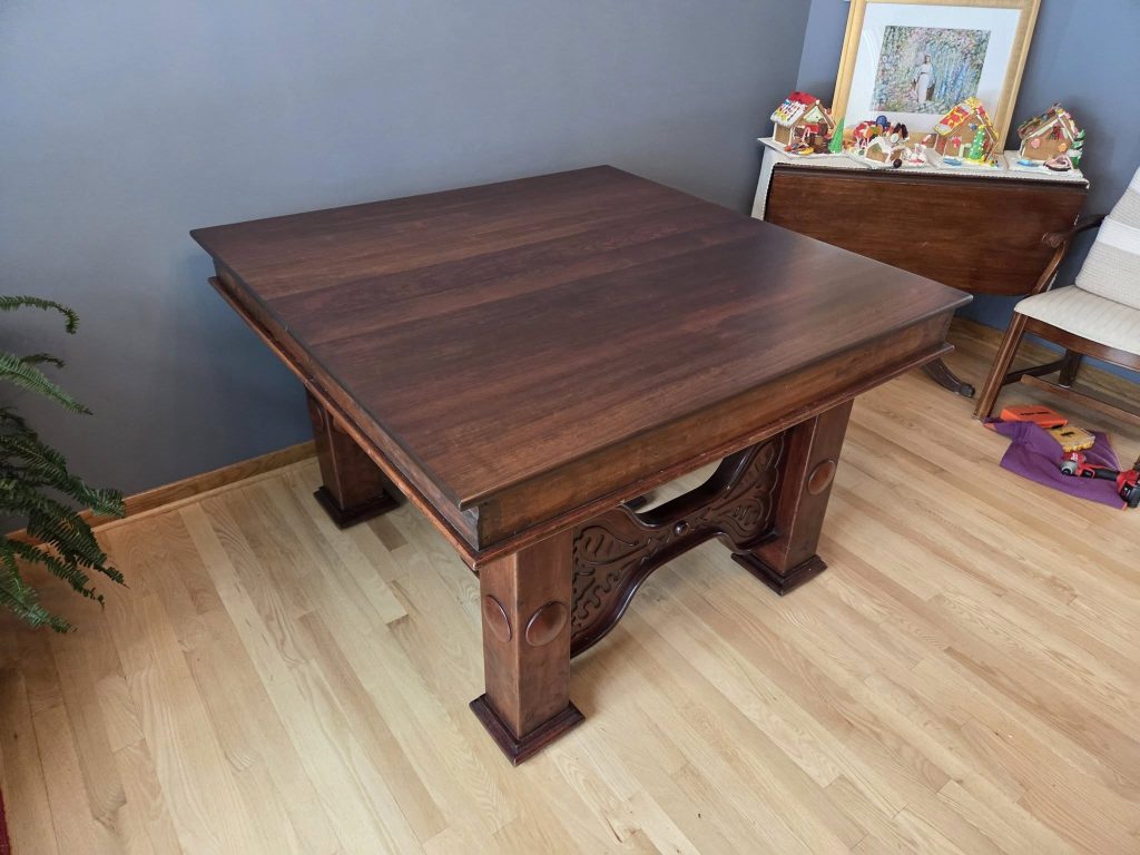 Dark wooden dining table with ornate carved legs in a blue-walled room; a sideboard holds gingerbread houses and a framed picture on top, with a chair nearby.