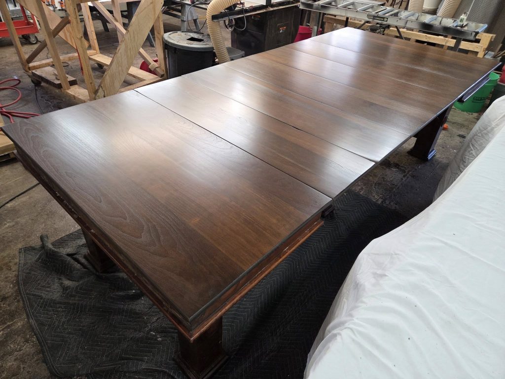 Long dark-stained wooden tabletop panels laid out in a woodworking shop for finishing, with tools and equipment in the background.
