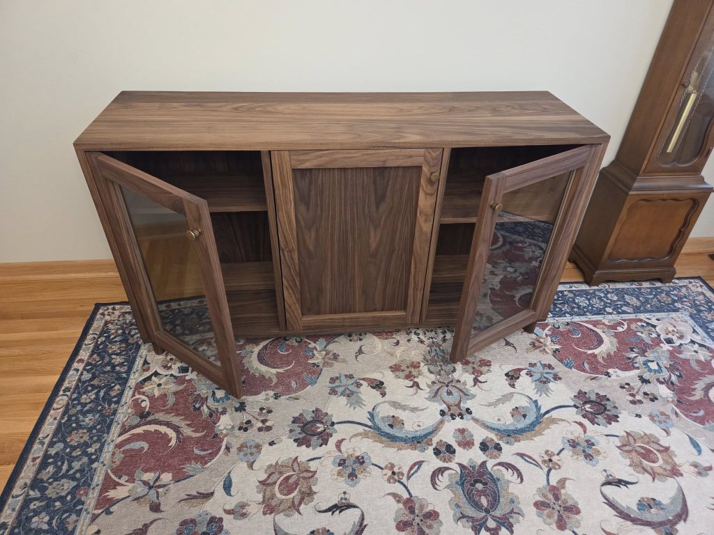 Wooden mid-century cabinet with glass doors open on both sides, central closed panel, in a room with a patterned rug and a grandfather clock nearby.