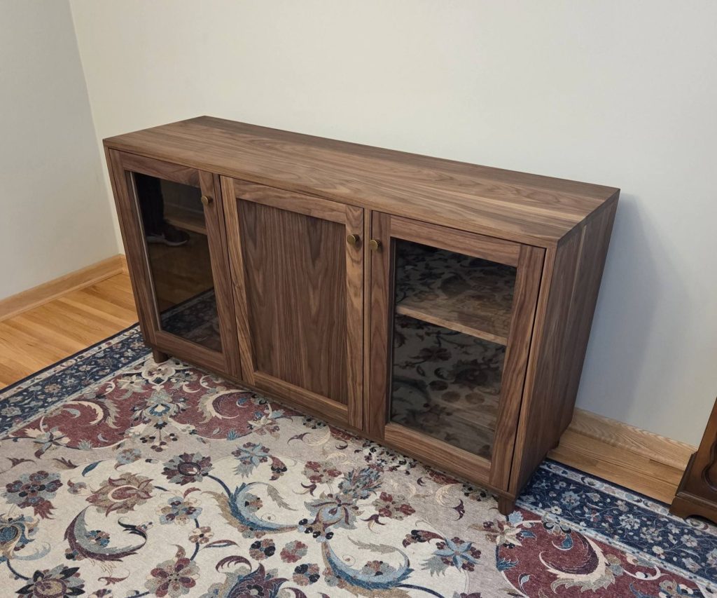 Wooden cabinet with glass doors and brass knobs, standing on a floral rug in a corner of a room.