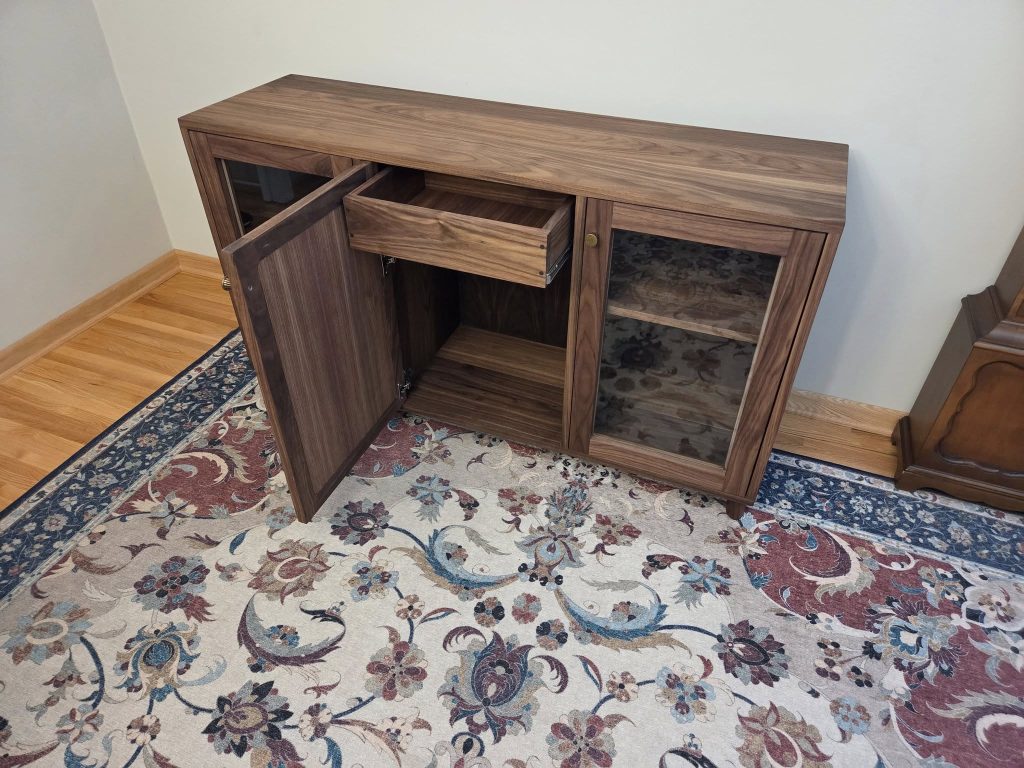 Wooden cabinet with two glass-front doors and an open drawer, showing interior shelves and storage space in a room with a patterned rug.