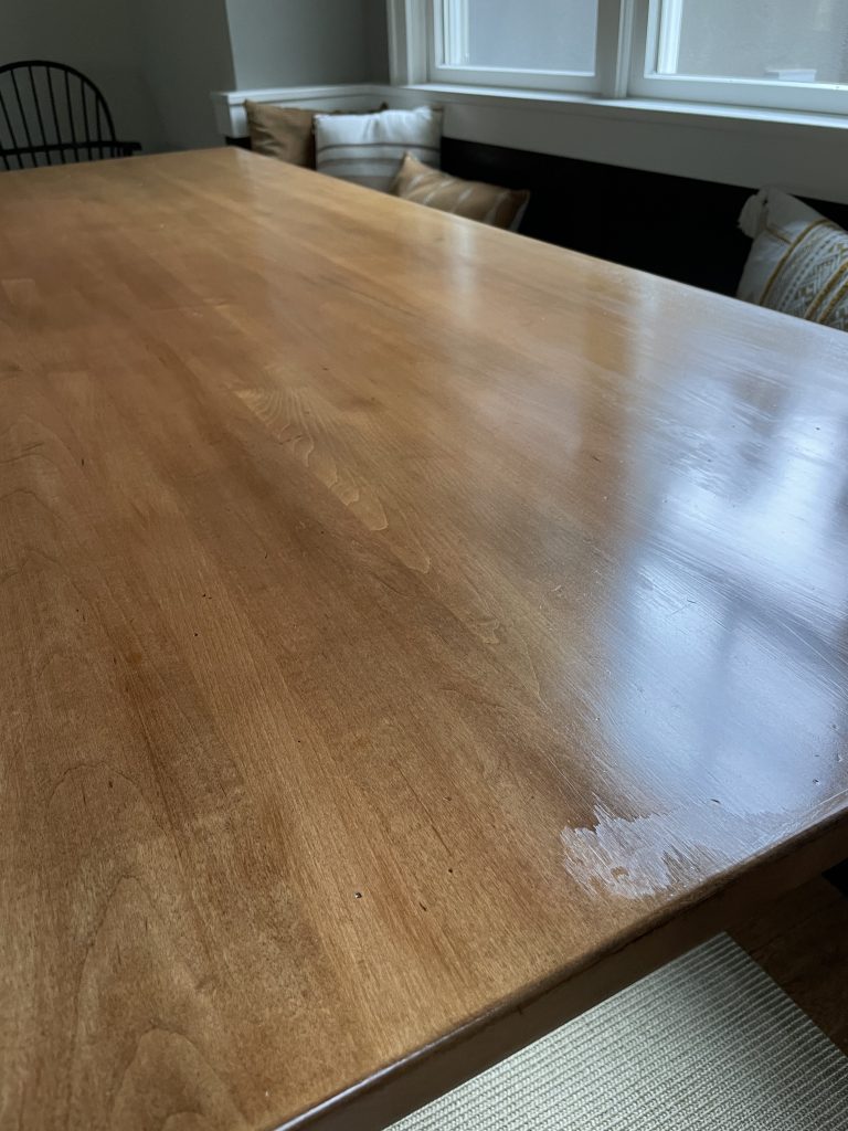 Wooden dining table with visible scratches and grain; cushions on a bench and a chair in the background by a window.