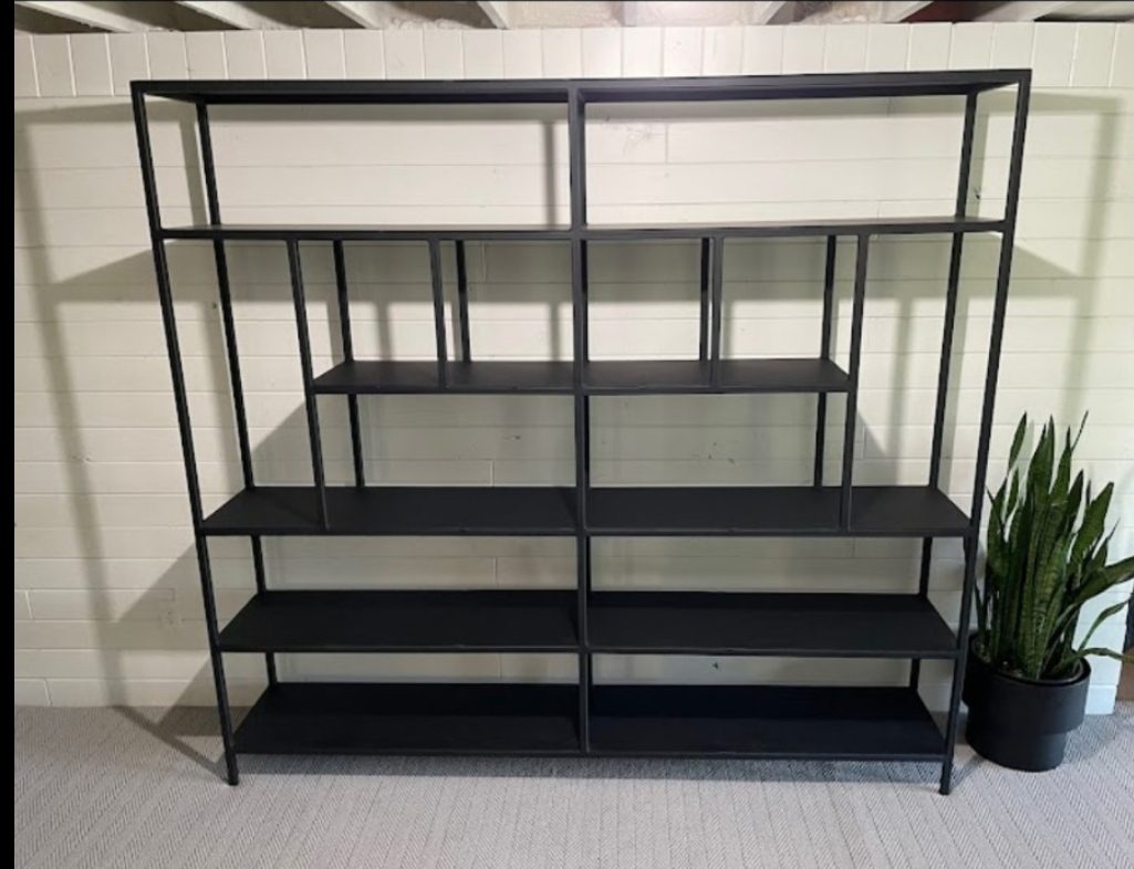 Black metal shelving unit with multiple open shelves, empty, placed against a white paneled wall; a tall potted plant sits to the right.