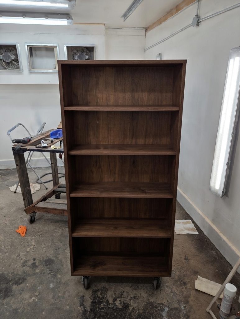 Tall dark wood bookcase with multiple shelves on caster wheels in a workshop, empty and ready for use.